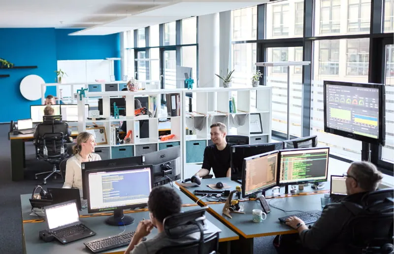 Mitarbeitende arbeiten in einem modernen Büro-Raum mit mehreren großen Bildschirmen voller System- und Netzwerkdaten.