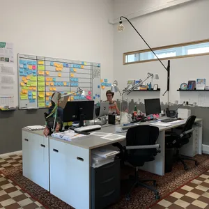 Zwei Mitarbeiterinnen arbeiten nebeneinander an ihren Schreibtischen. An der Wand hinter ihnen hängen viele bunte Post-its.