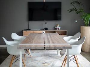 Ein Holztisch und Stühle stehen in einem Meetingraum, an der Wand hängt ein großer Bildschirm.
