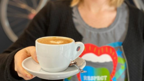 Eine Mitarbeiterin hält eine Tasse Kaffee mit einem Herz aus Milchschaum in die Kamera.